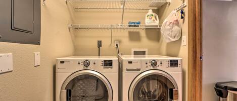 A laundry area with two front-loading washing machines, a metal shelf with laundry supplies, and a trash can.