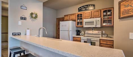 A kitchen with a large island, wooden cabinets, a microwave, and a refrigerator. There are bar stools at the island and a sign on the wall that says "The Best Place to Be is Together".