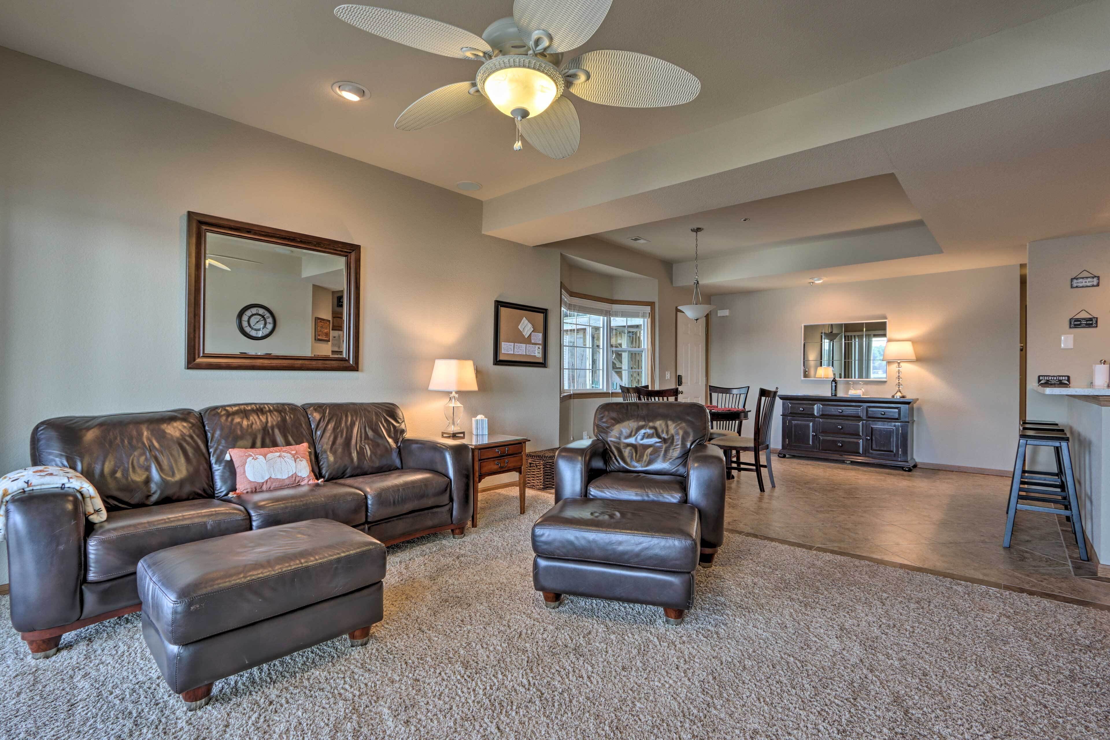 A spacious living room with a large leather sofa, ottoman, and a ceiling fan. There is a dining area with a table and chairs, and a kitchen in the background.