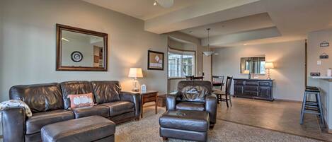 A spacious living room with a large leather sofa, ottoman, and a ceiling fan. There is a dining area with a table and chairs, and a kitchen in the background.