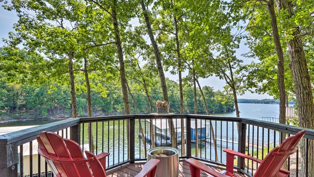 A deck with red Adirondack chairs, a metal fire pit, and a view of a lake with boats.