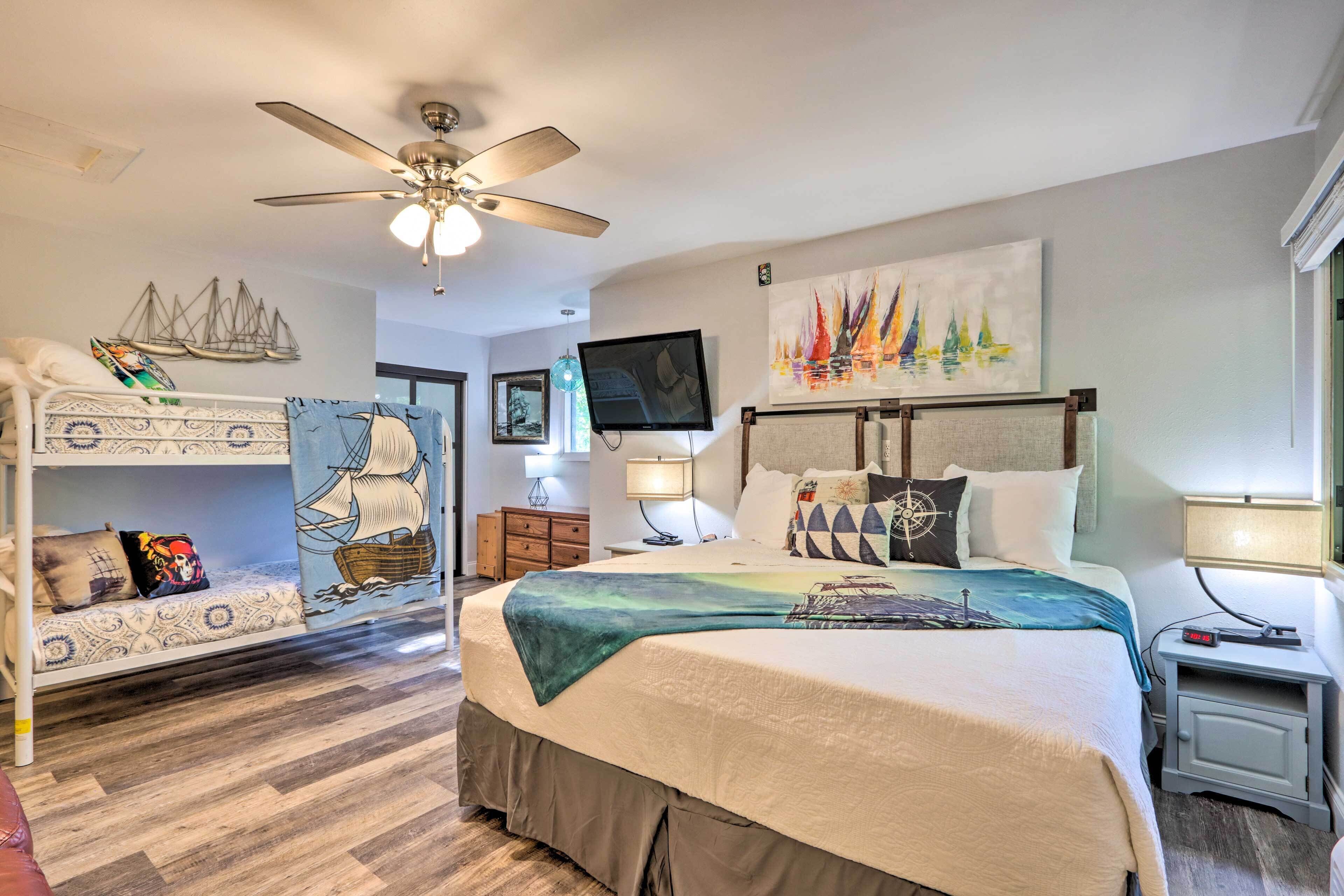 A hotel room with a bed, a ceiling fan, a TV, and a painting of sailboats.