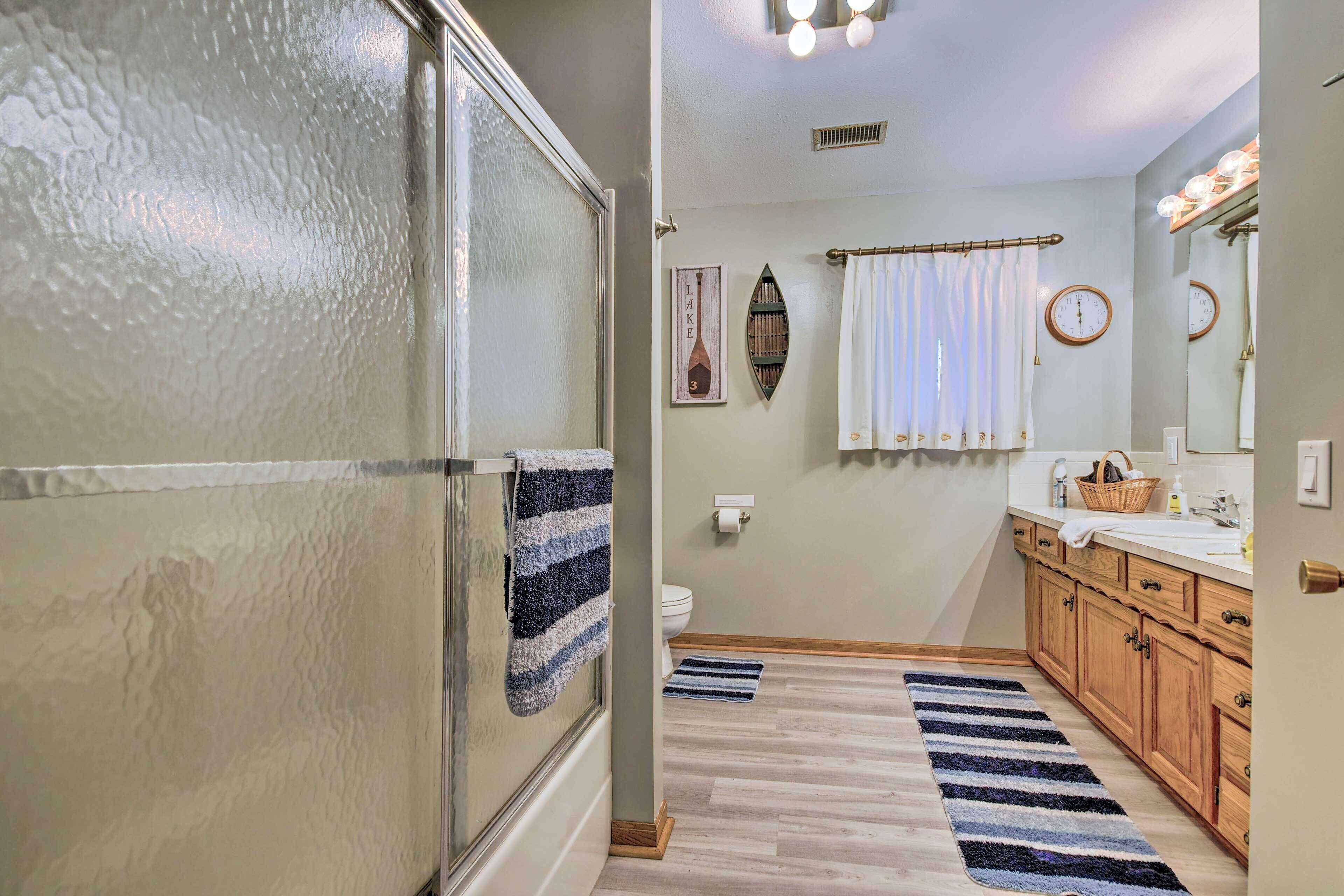 A bathroom with a glass shower door, wooden cabinets, a white sink, and a striped rug.