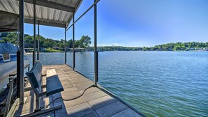 A covered dock with a bench, a boat, and a view of a lake with buildings and trees in the distance. - Boat Dock + Slides: Lakefront Gravois Mills Home (Gravois Mills)