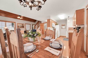 A dining area with a wooden table set for four, surrounded by wooden chairs. A chandelier hangs from the ceiling, and there is a kitchen area visible in the background.