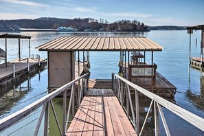 A wooden dock with a covered seating area and a small building, overlooking a calm lake with a distant shoreline. - Lakefront Ozarks Home: Deck + Boat Dock! (Linn Creek)
