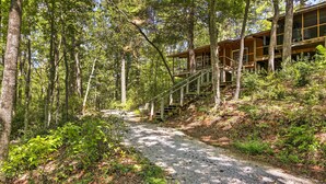 Zona boscosa con un sendero de grava que lleva a una cabaña de madera con escaleras.