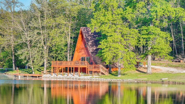 Une cabane dotée d’un toit triangulaire, d’une terrasse en bois et d’un quai s’avançant dans un lac, le tout entouré d’arbres.