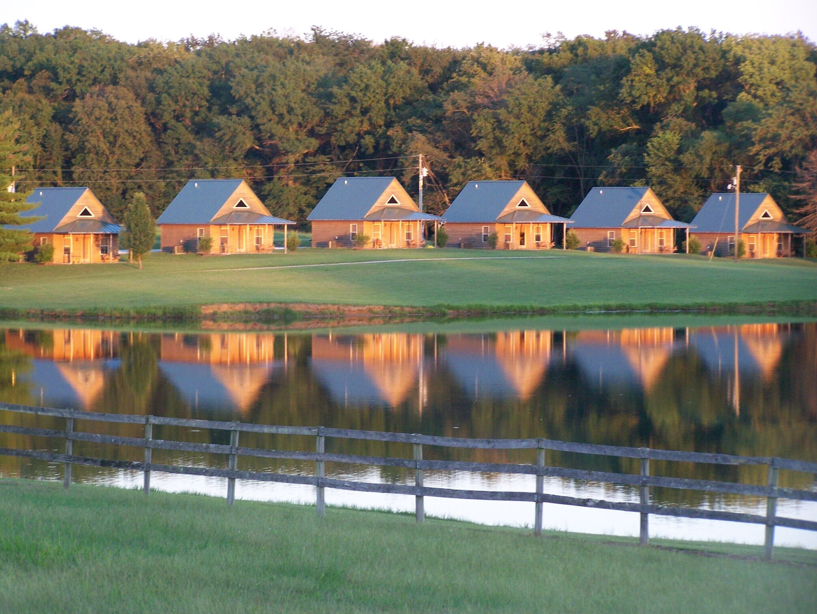 A row of cabins with a lake in the foreground and a forested hill in the background.