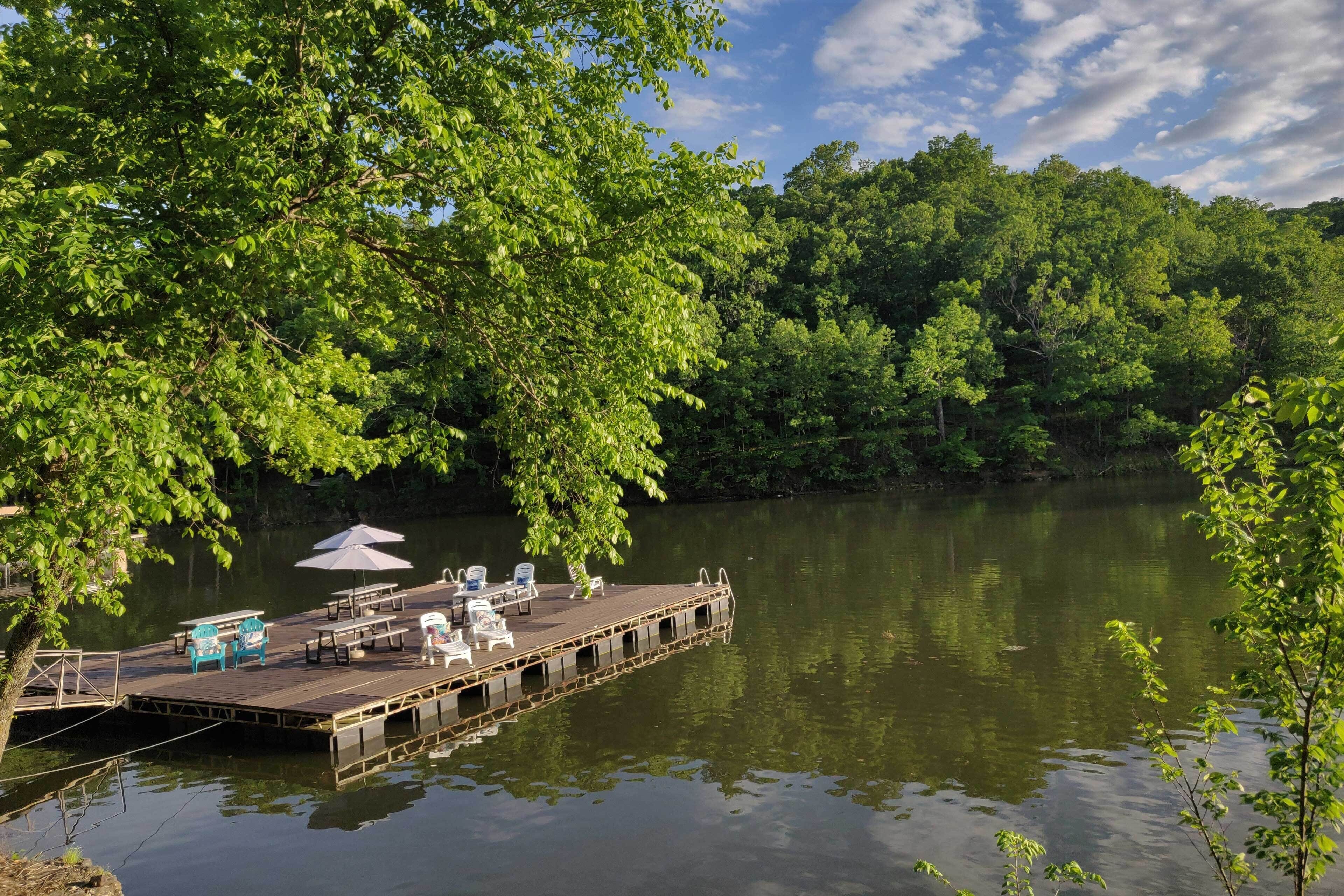 A dock with chairs and tables, surrounded by lush greenery and a calm lake.
