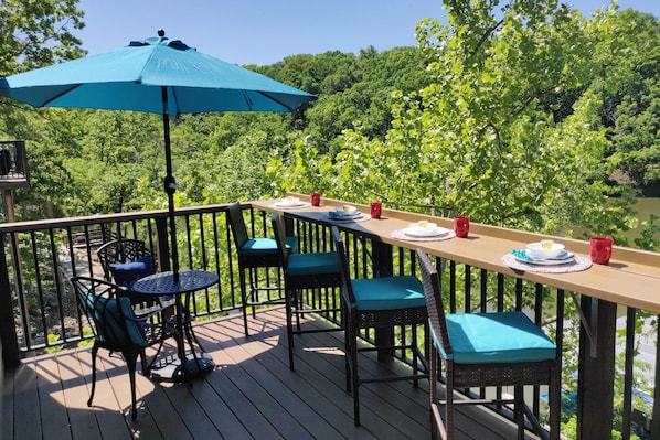 A wooden deck with a table and chairs, a blue umbrella, and a view of a forested area.