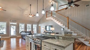 A spacious kitchen with granite countertops, a built-in oven, and a large island. There is a wooden staircase with a chandelier and ceiling fan. - Lakefront Retreat w/ Game Room & Private Dock! (Bonneau)