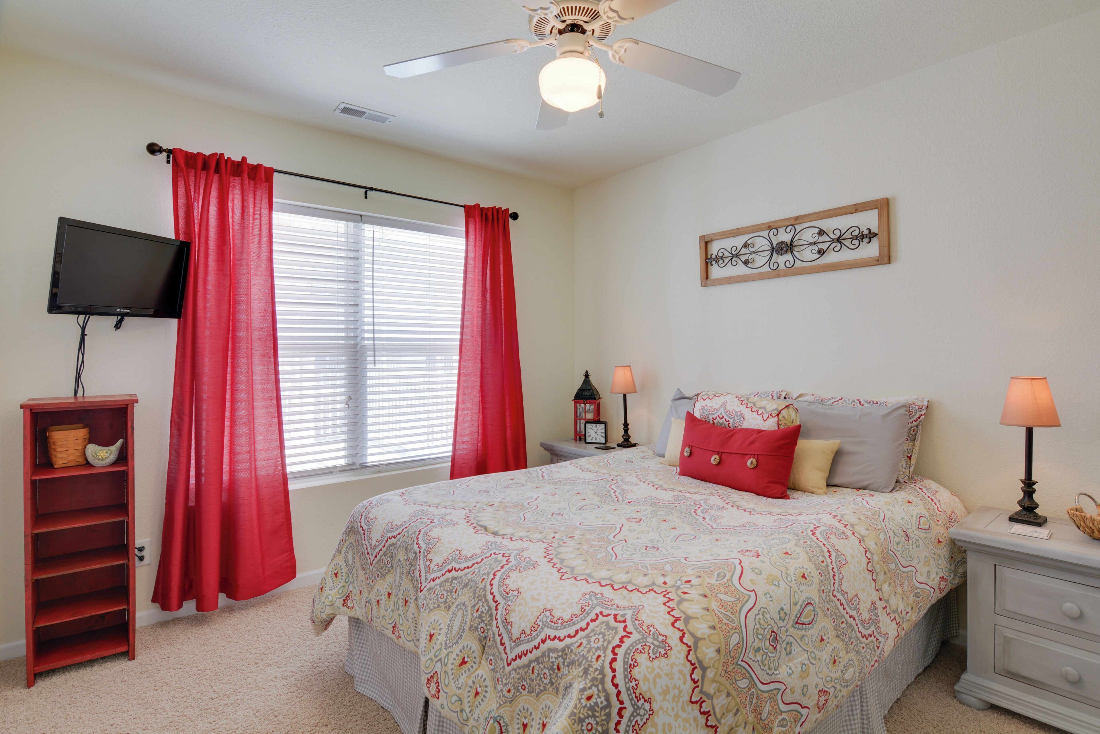 Quarto com cama, televisão, mesa de cabeceira, abajur, estante e janela com cortinas.