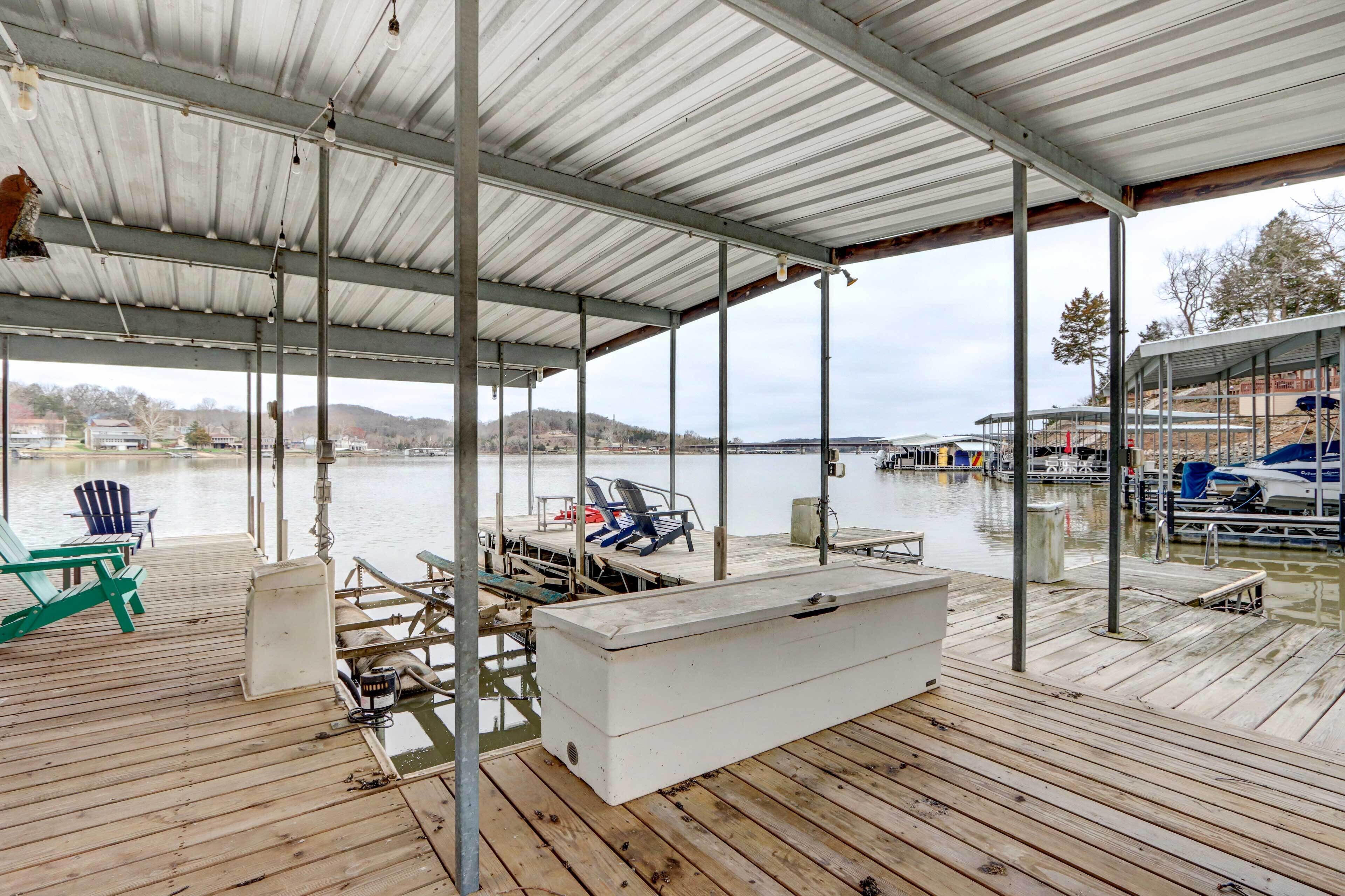 Un muelle techado con sillas y un enfriador, con vista a una masa de agua tranquila y barcos al fondo.