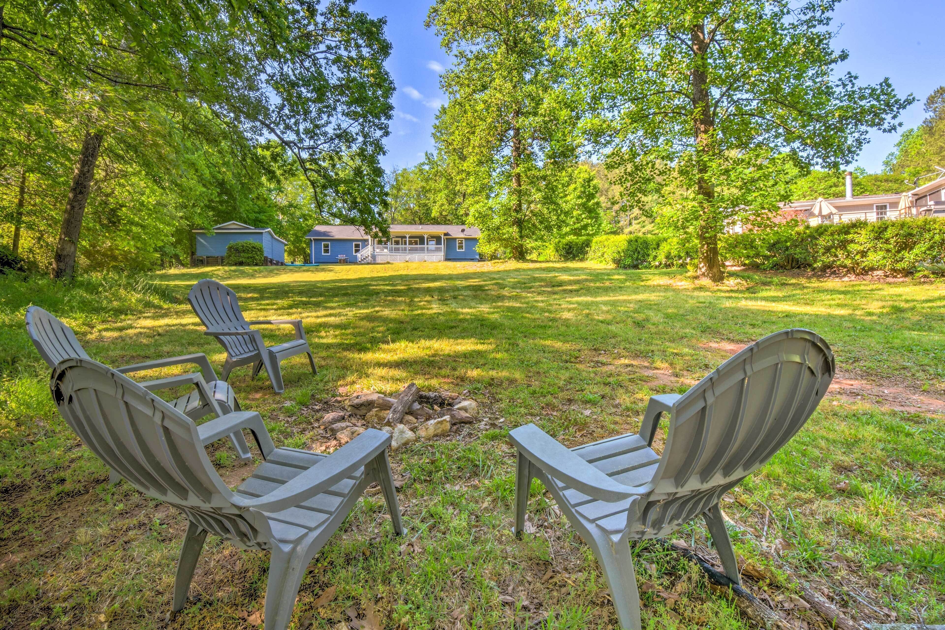 A backyard with two Adirondack chairs facing a stone fire pit, surrounded by lush greenery and a house in the background.