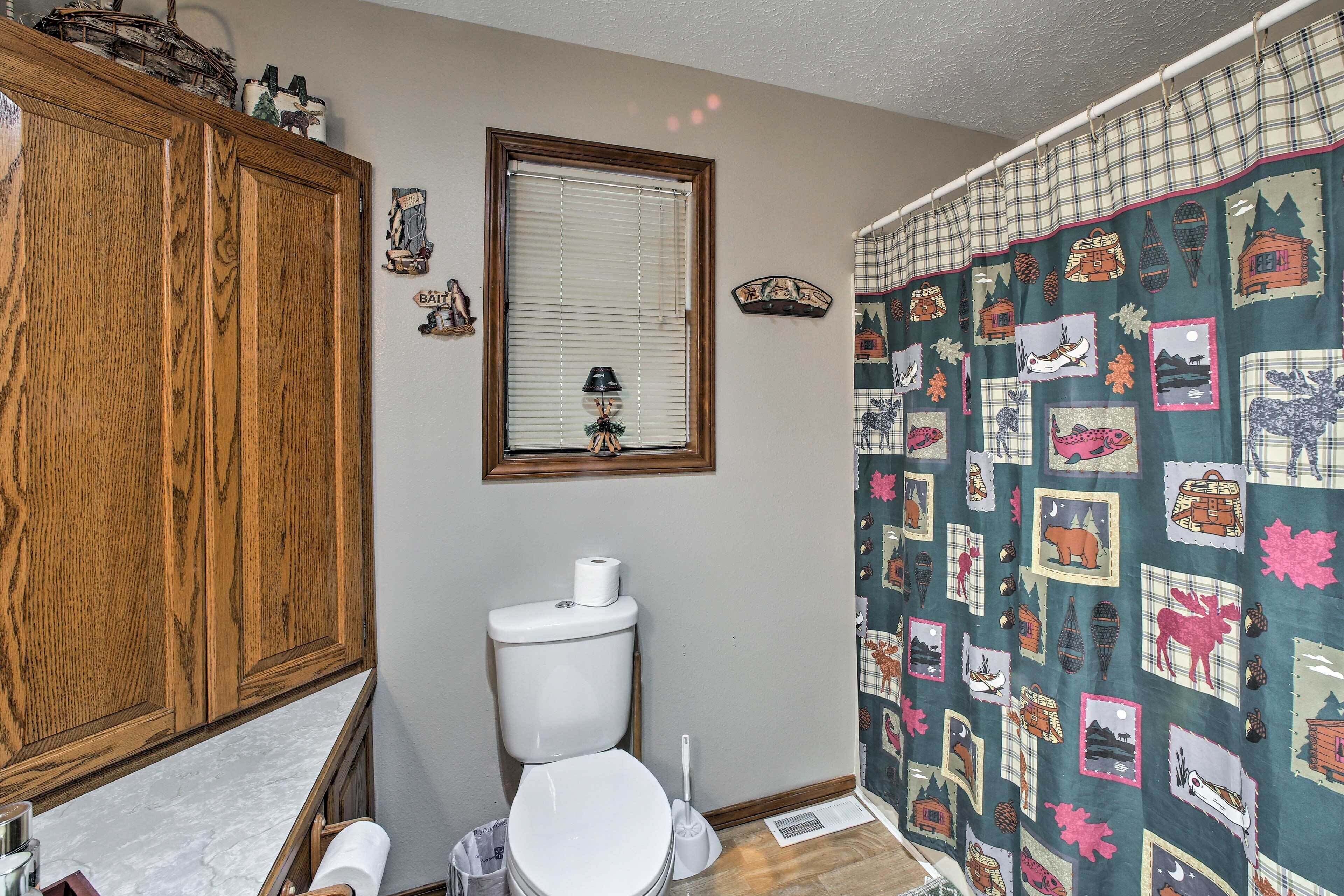 A bathroom with a wooden cabinet, a toilet, a window with blinds, and a shower curtain with a forest theme.