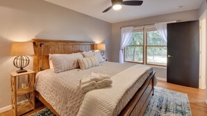 Un dormitorio con cama, ventilador de techo, mesita de noche con lámpara y ventana con cortinas.