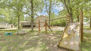 A playground with a swing set and a slide, surrounded by trees and a house. - Lakeside Reeds Spring Home w/ Deck & Pool Access! (Kimberling City)