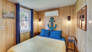 A wooden-paneled room with a bed, a wall-mounted air conditioner, a small table with a lamp, and a window with curtains. - Lakeside Reeds Spring Home w/ Deck & Pool Access! (Kimberling City)