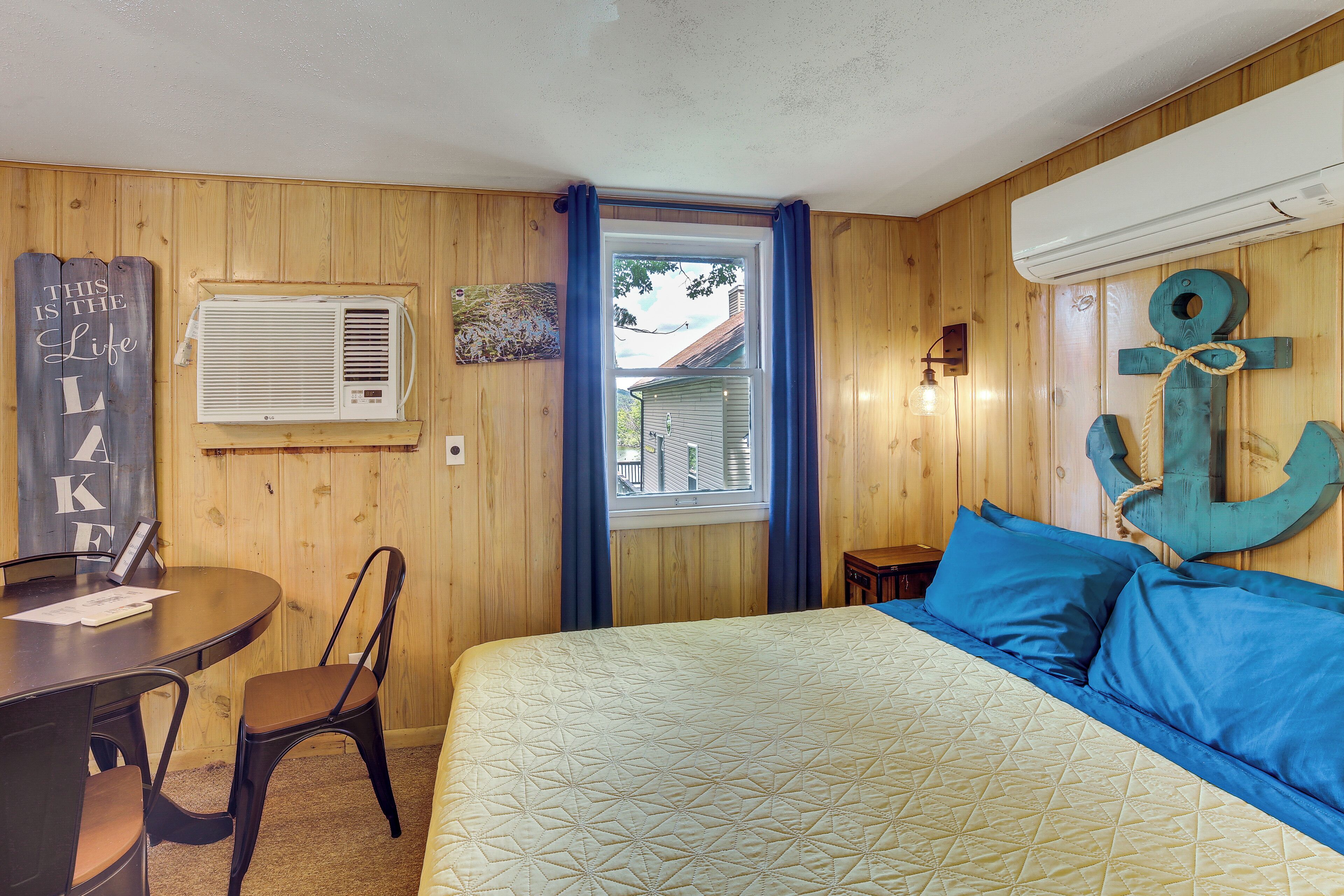 A room with a bed, a table with chairs, an air conditioning unit, a window with curtains, and a wall-mounted anchor decoration.