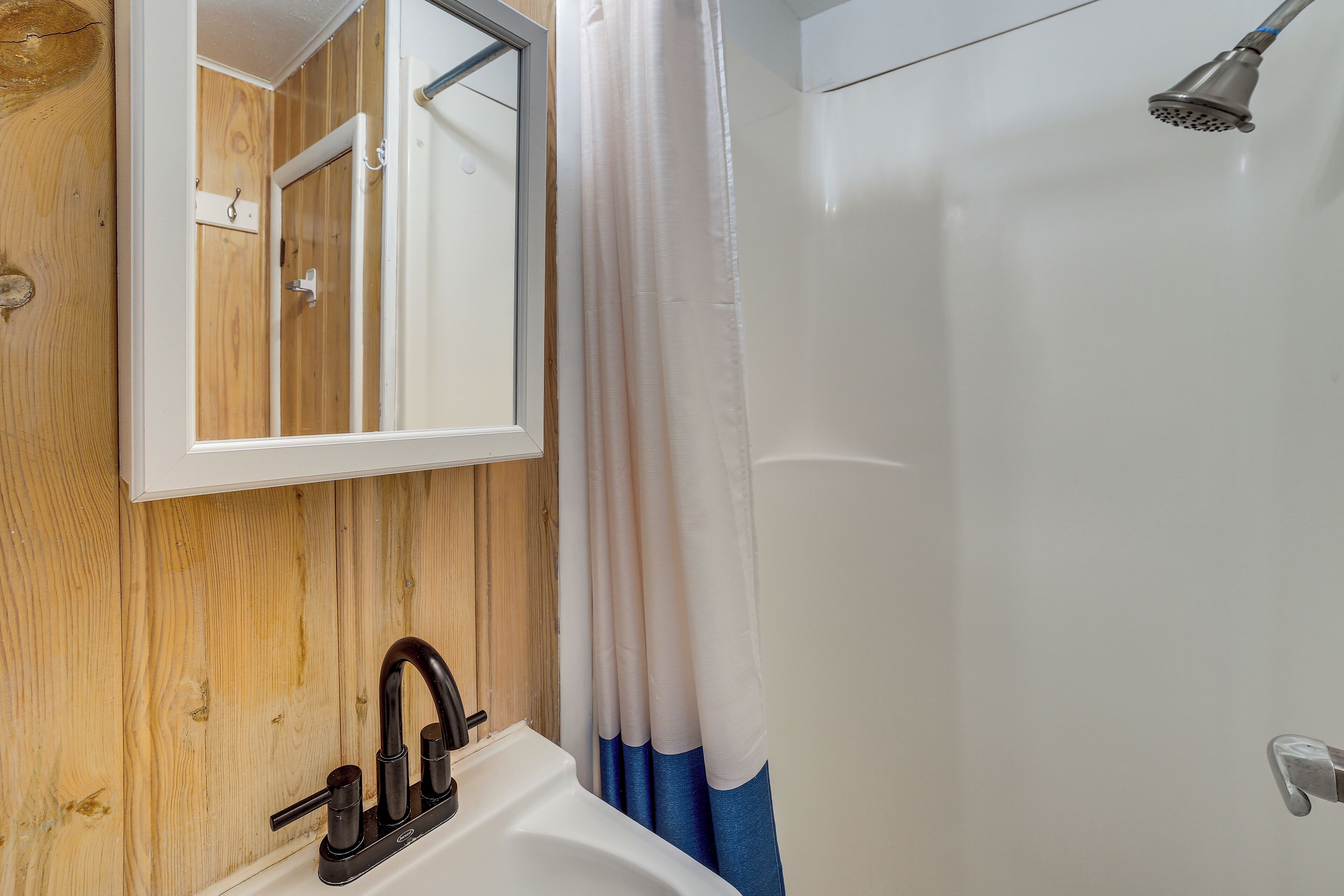 A bathroom with a wooden wall, a white sink, a black faucet, a shower with a white curtain, and a mirror.