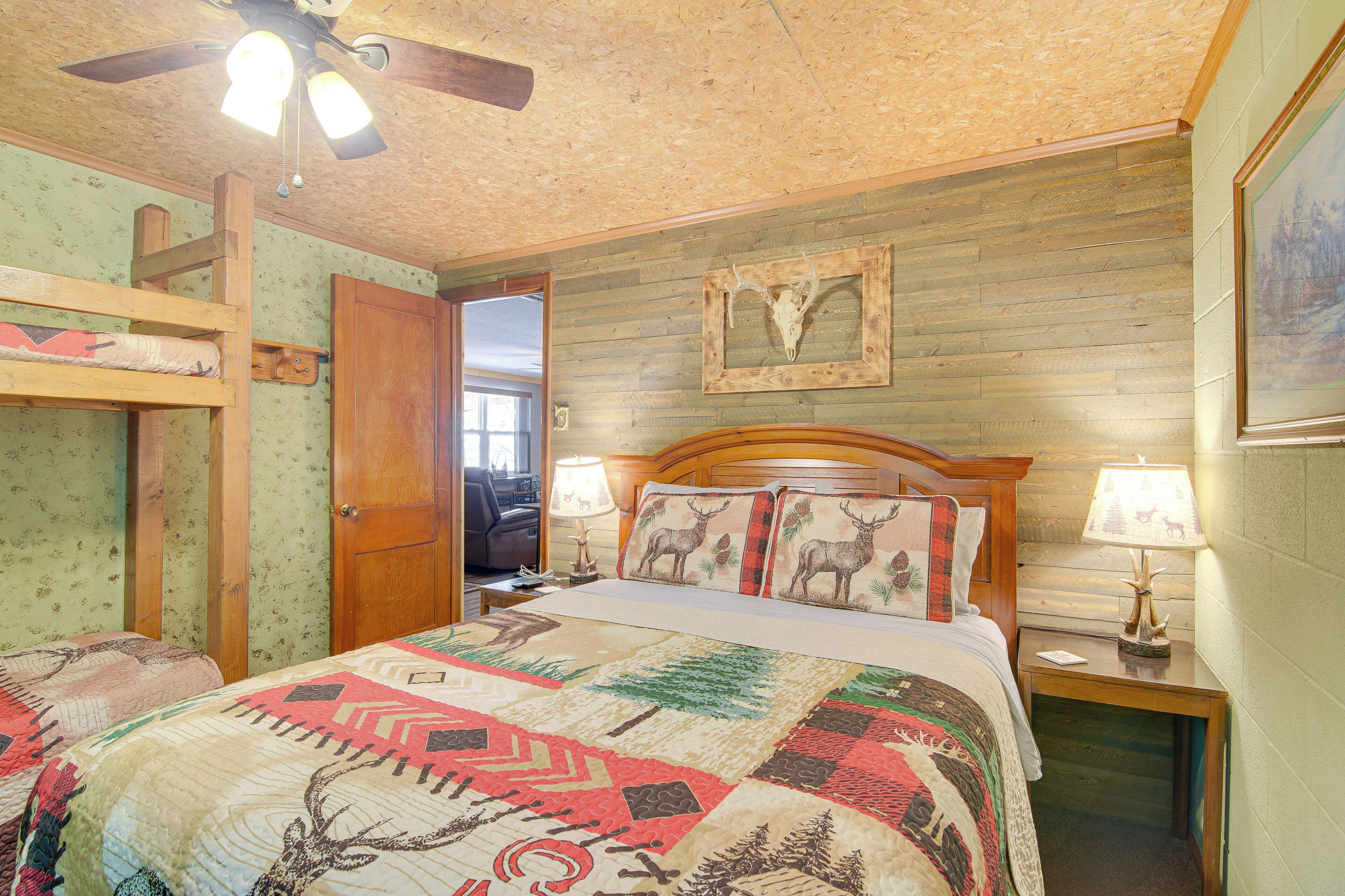 A rustic bedroom with a wooden ceiling, a bunk bed, a large bed with a quilt, a wooden headboard, two bedside lamps, and a framed deer skull on the wall.