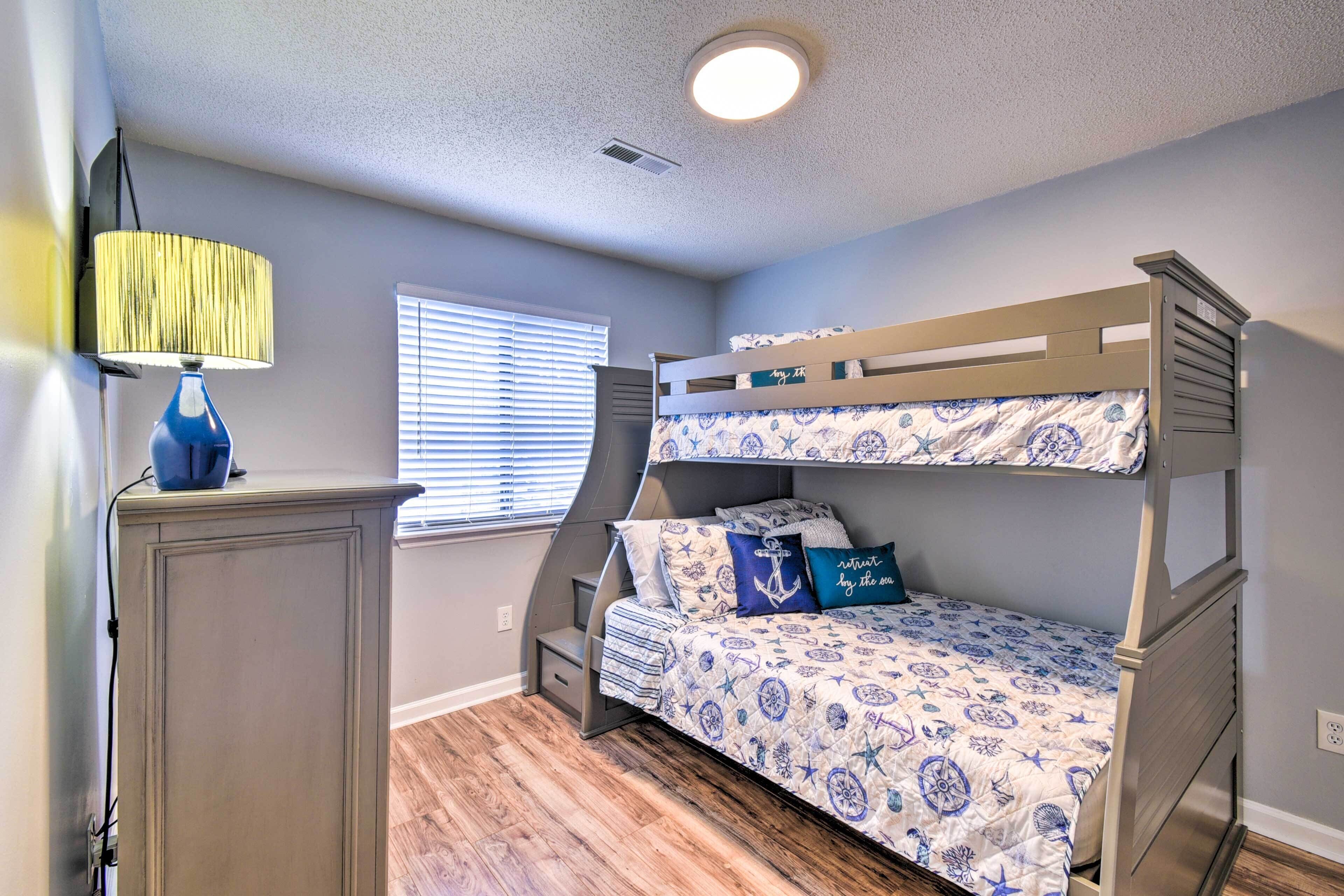 A bunk bed with a nautical-themed bedspread, a desk with a lamp, and a window with blinds.