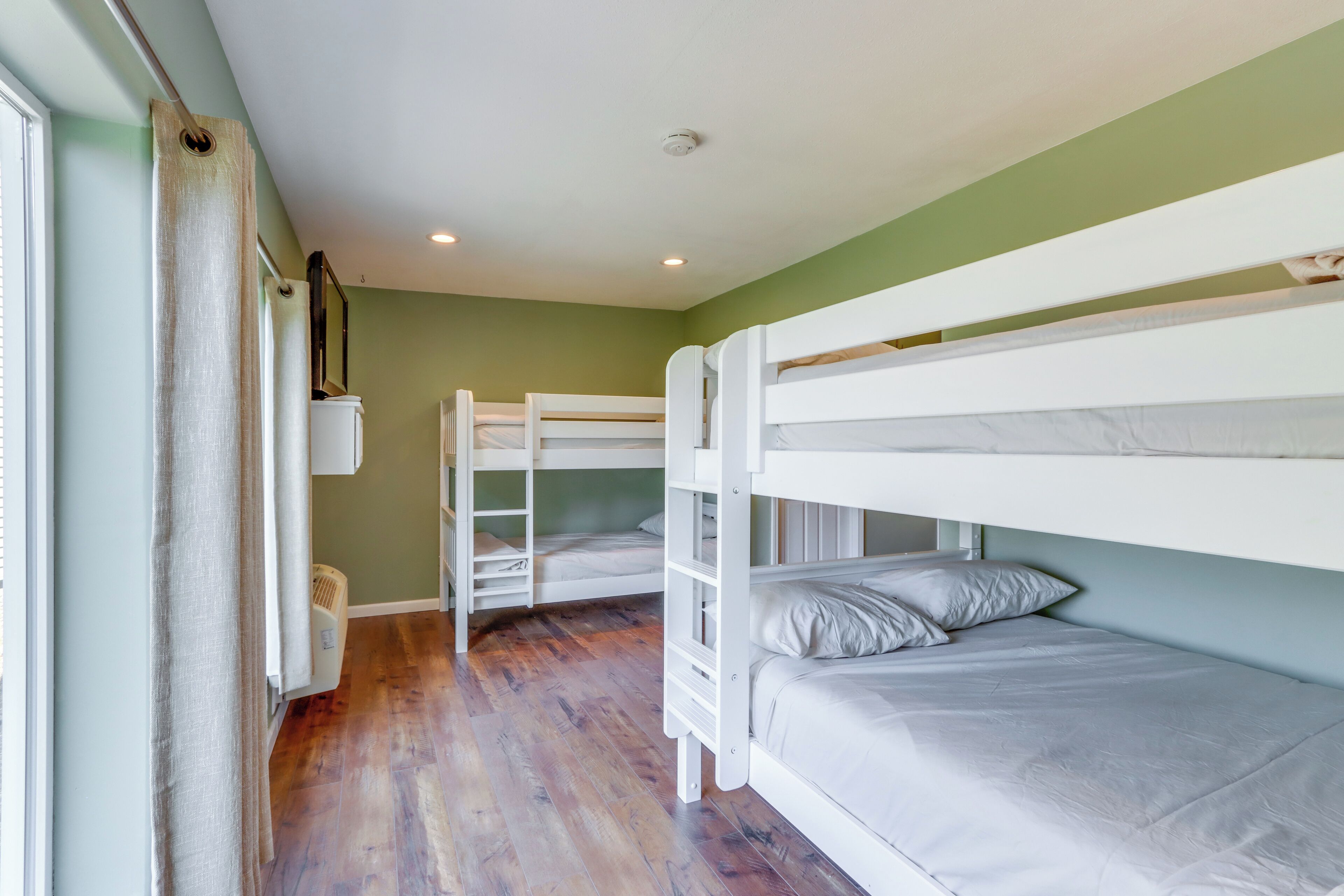 Dormitorio con literas, suelo de madera, radiador y ventana con cortinas.