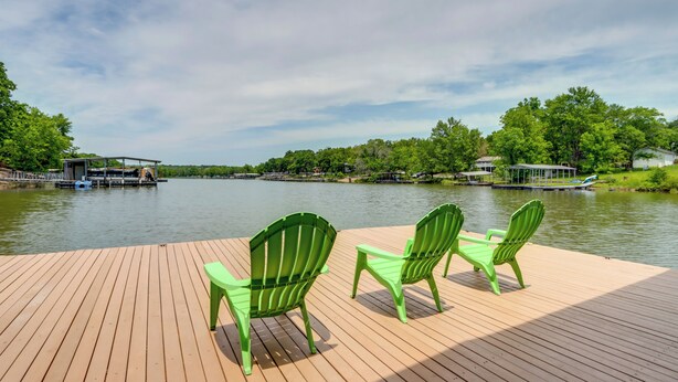 A wooden deck with three green Adirondack chairs facing a calm lake, surrounded by lush greenery and a dock with boats. - 'lazy RS Lakehouse' w/ Hot Tub & Dock in Warsaw! (Warsaw)
