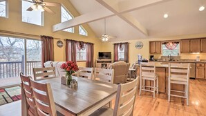 A spacious living area with a dining table, chairs, a sofa, and a TV. There are large windows with curtains, a ceiling fan, and a kitchen area with a sink and cabinets. - 'lazy RS Lakehouse' w/ Hot Tub & Dock in Warsaw! (Warsaw)