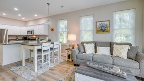 A modern kitchen with white cabinets, a dining area with a white table and chairs, a gray sofa with white cushions, and a wooden coffee table.