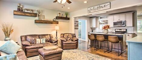 A modern living room with a leather sofa, ottoman, and a coffee table. There is a kitchen area with a bar and stools, a ceiling fan, and a 'Welcome' sign.