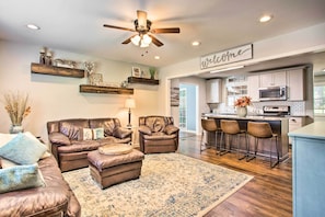 A modern living room with a leather sofa, ottoman, and a coffee table. There is a kitchen area with a bar and stools, a ceiling fan, and a 'Welcome' sign.
