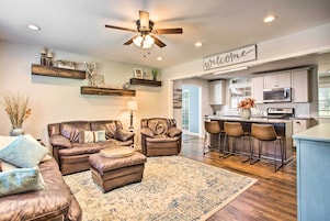 A modern living room with a leather sofa, ottoman, and a coffee table. There is a kitchen area with a bar and stools, a ceiling fan, and a 'Welcome' sign.