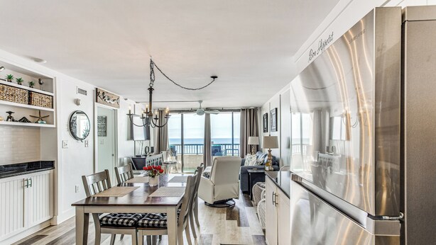 A modern kitchen with a large stainless steel refrigerator, a dining area with a wooden table and chairs, and a view of the ocean through large windows. - Oceanfront Oasis in North Myrtle Beach (North Myrtle Beach)