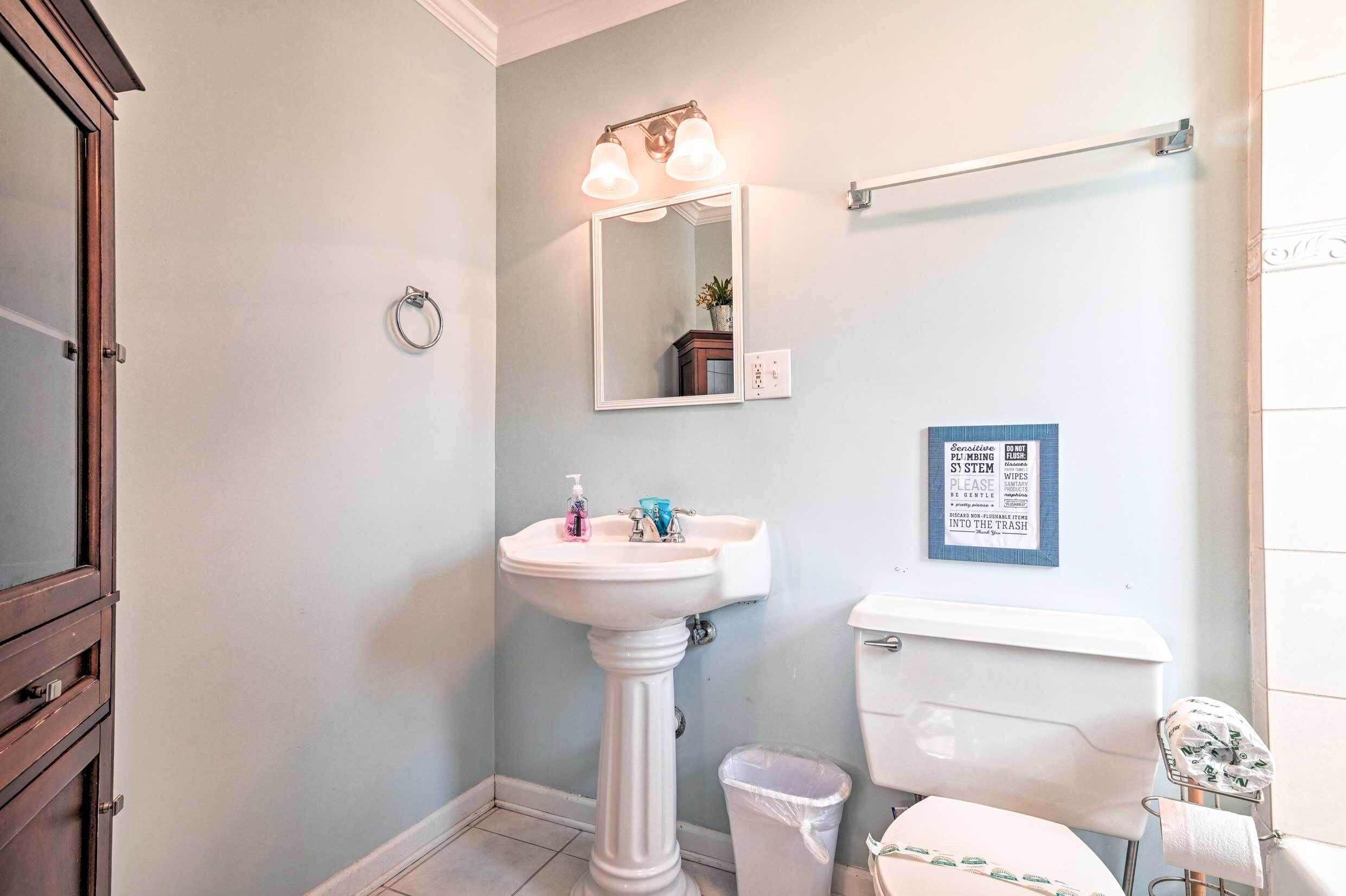 A bathroom with a pedestal sink, a mirror, a toilet, and a trash bin.