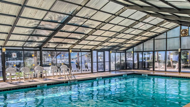 An indoor swimming pool with clear glass walls, a metal roof, and a poolside area with chairs and a ladder. - 1 Mi to Beach + Hot Tub: Murrells Inlet Retreat! (Murrells Inlet)