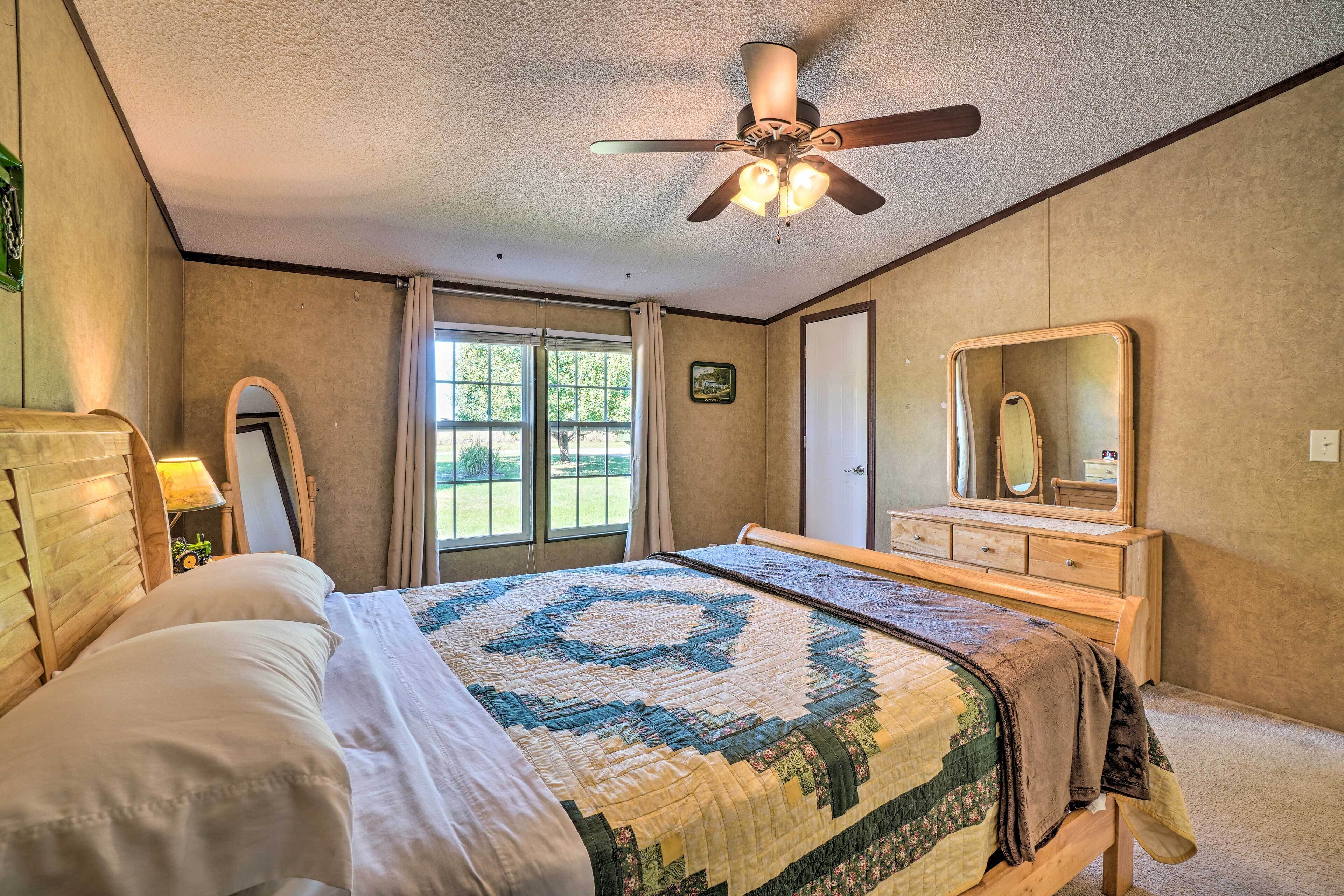 Une chambre à coucher comprenant un lit, un ventilateur de plafond, une commode, un miroir et une fenêtre avec des rideaux.