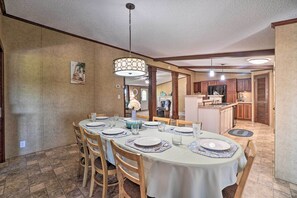 A dining area with a round table set for six, surrounded by wooden chairs. A chandelier hangs from the ceiling, and a kitchen is visible in the background.
