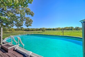 A clear blue swimming pool with wooden deck and metal railings, surrounded by a fenced grassy area and trees.