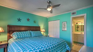 Ein Schlafzimmer mit einem Bett, einem Deckenventilator, einem Nachttisch, einer Lampe und einer Tür zu einem Badezimmer.