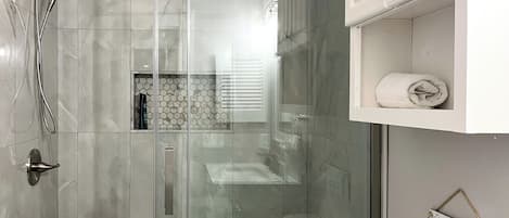 A modern bathroom with a glass shower enclosure, a white towel on a shelf, and a sign requesting not to flush paper towels.
