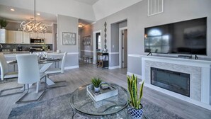 Un salon moderne avec une table basse en verre, une cheminée, un téléviseur à écran plat et un coin repas comprenant des chaises blanches et un lustre.