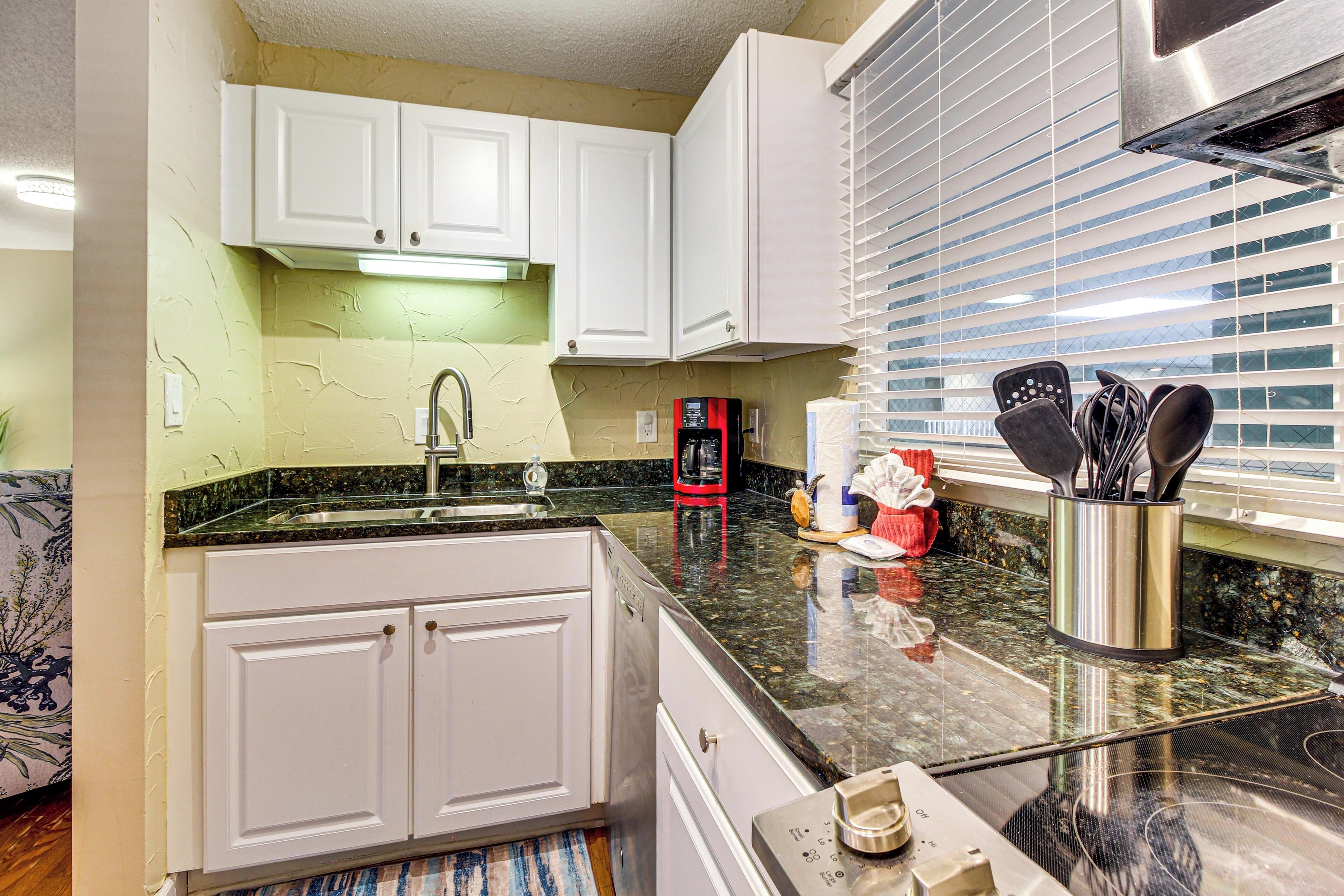 A kitchen with white cabinets, a stainless steel sink, a black countertop, and a window with blinds.