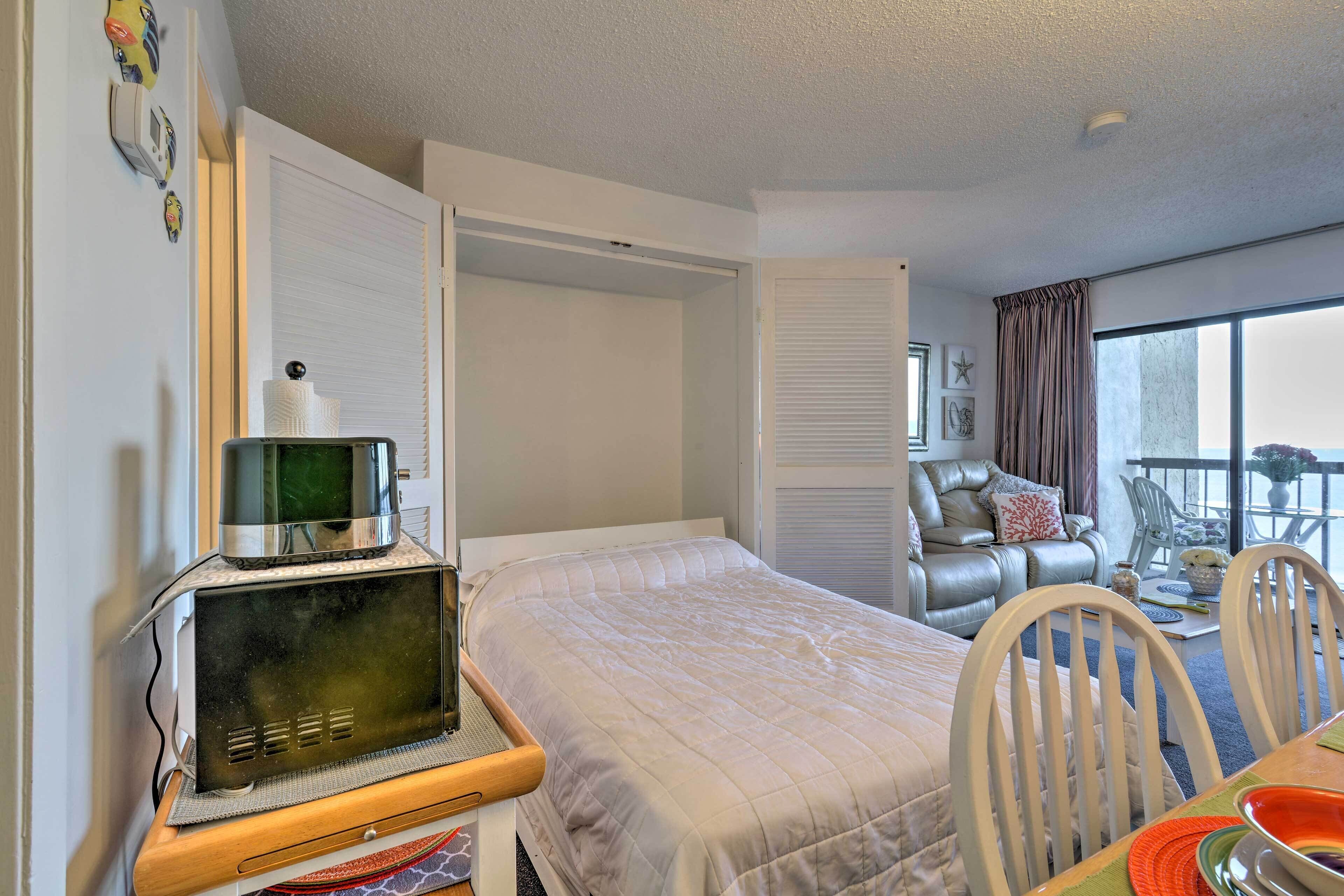 A hotel room with a bed, a microwave, a dining table, and a view of the outside.