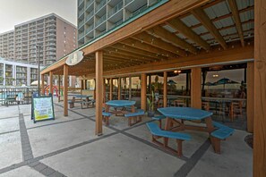 An outdoor patio area with wooden tables and benches, a covered seating area, and a view of a high-rise building in the background. - Walk to Ocean: Myrtle Beach Condo w/ Views! (Myrtle Beach)