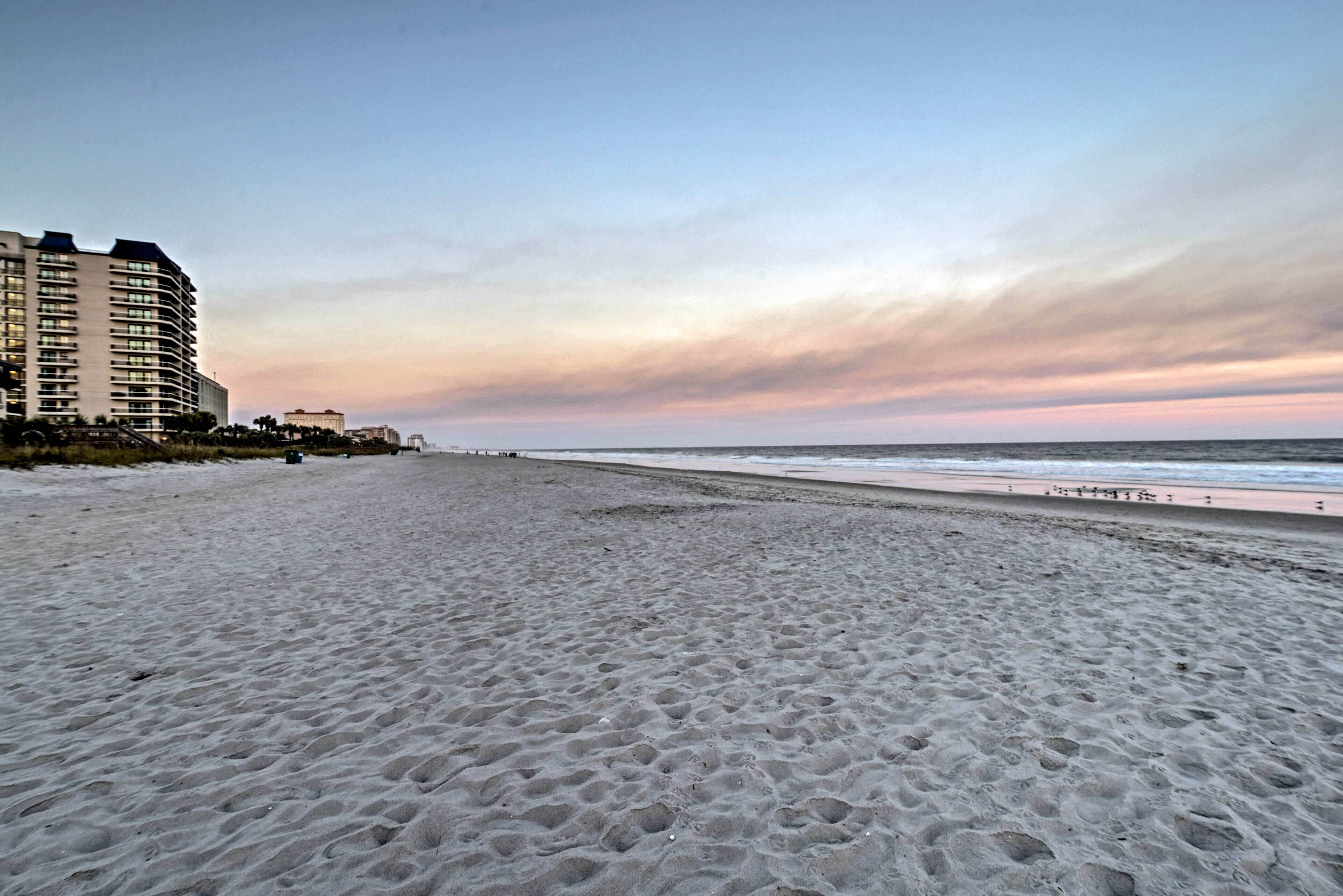 A beach with a sandy shore, a few buildings in the background, and a clear sky.