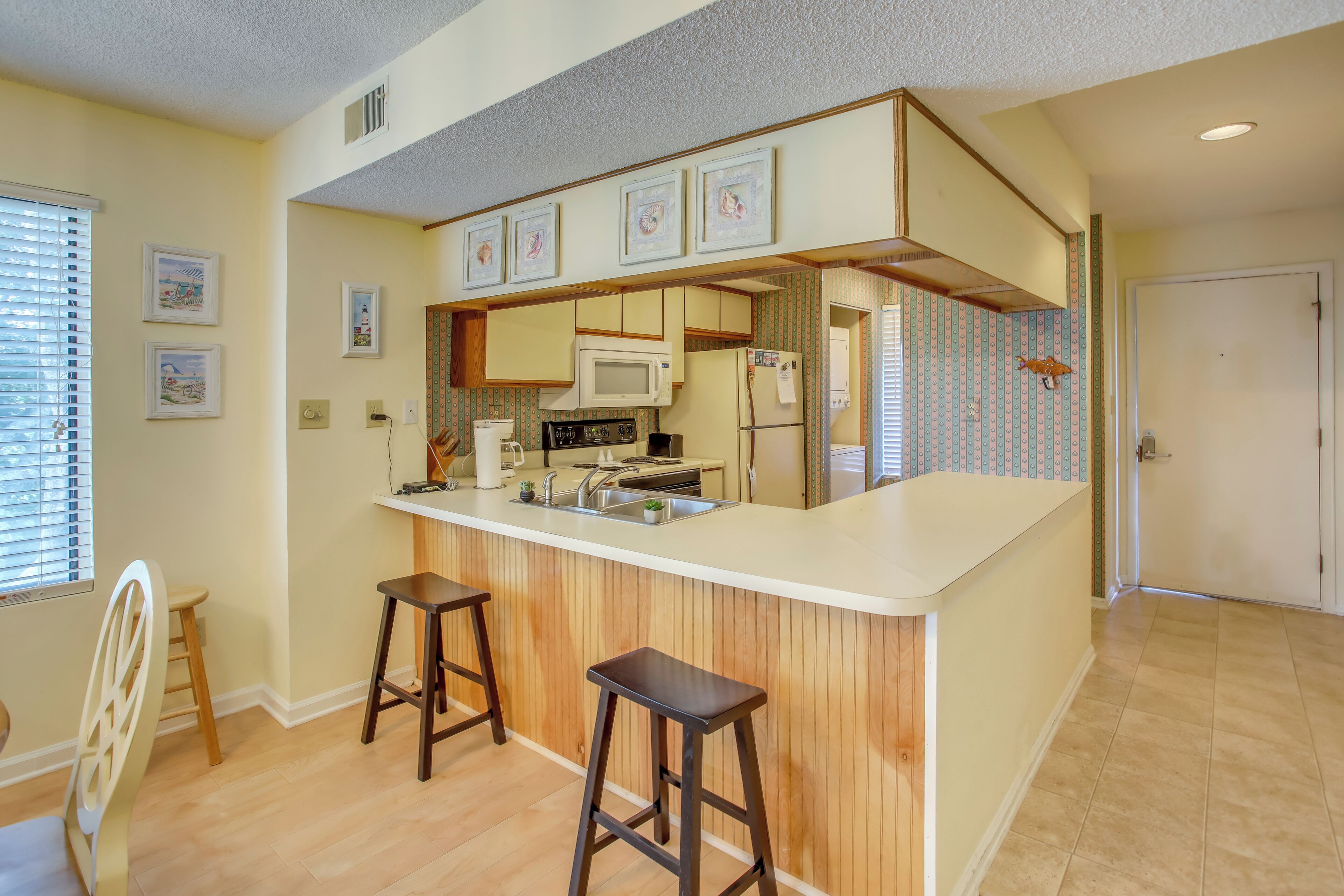 A kitchen with a white countertop, wooden cabinets, a microwave, and a refrigerator. There are two stools and a window with blinds.