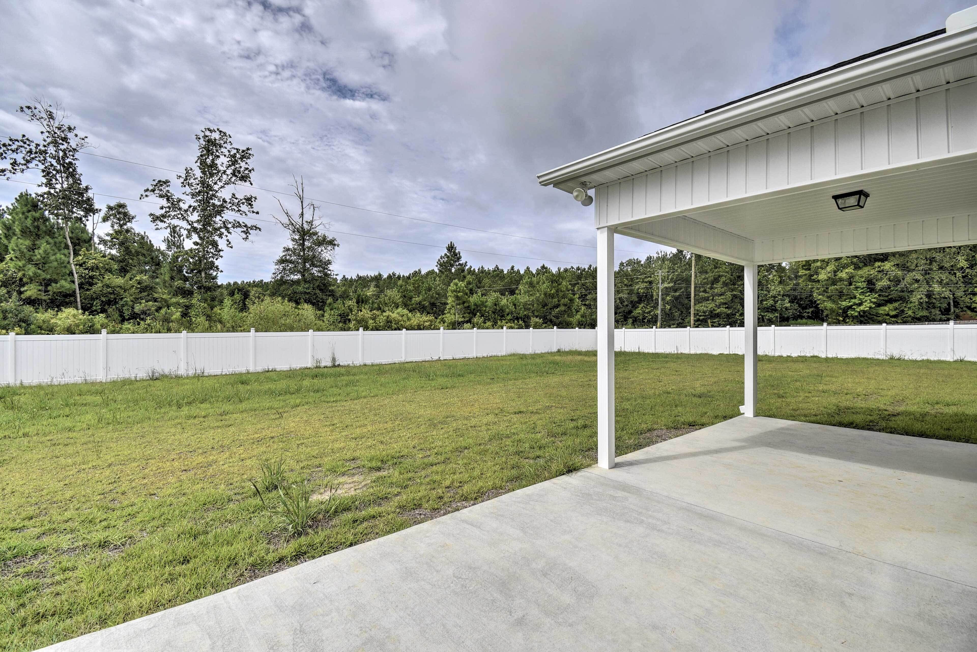 A covered patio area with a concrete floor, surrounded by a white fence and a grassy lawn.