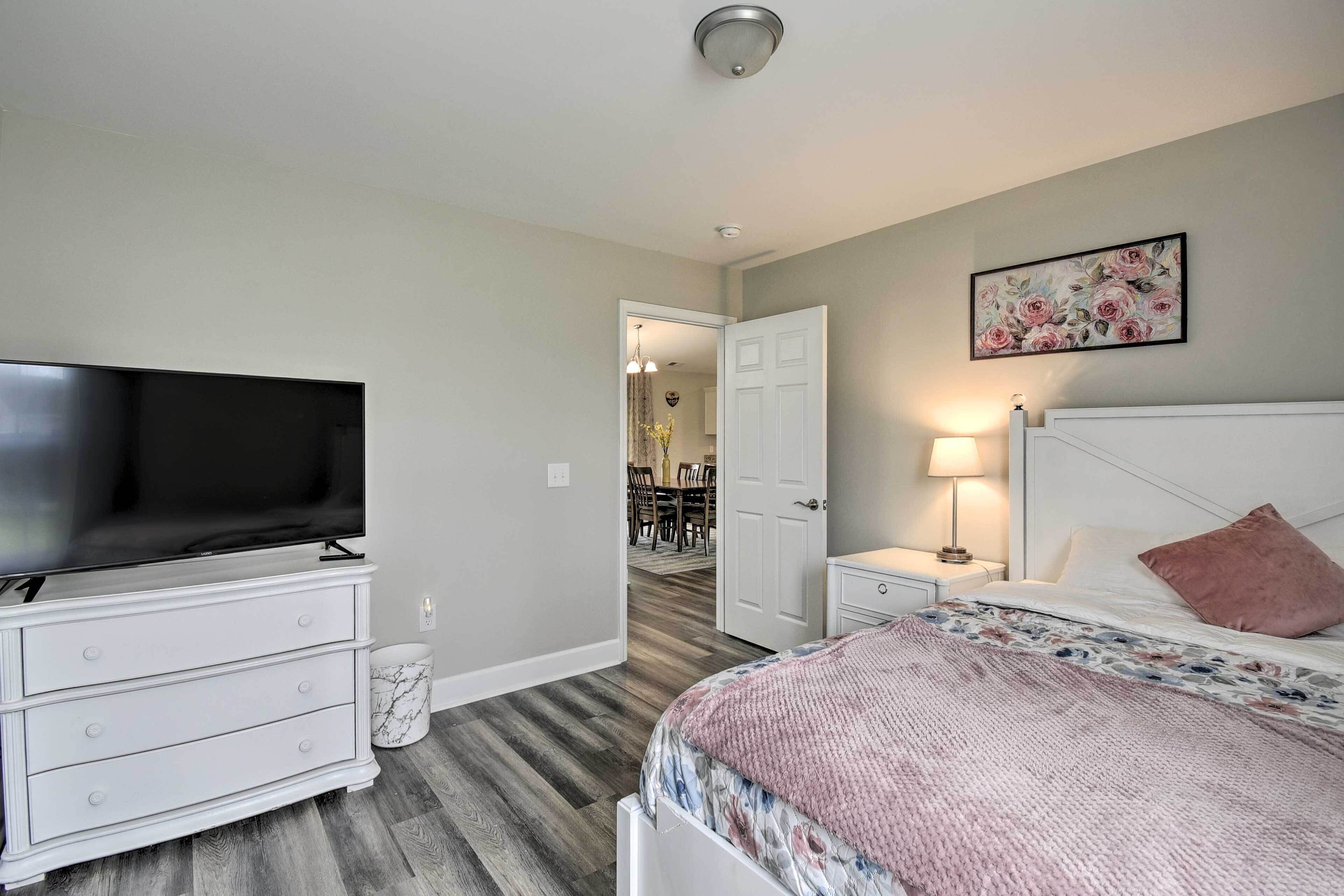 A bedroom with a TV, a bed with a floral bedspread, a nightstand with a lamp, and a door leading to a dining area.